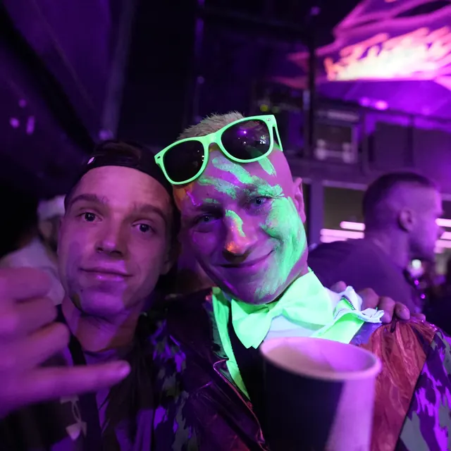 To festivalgæster i lilla lys, den ene med neongrøn ansigtsmaling og solbriller på hovedet.