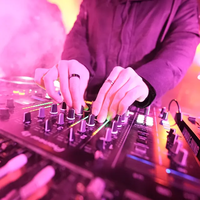 Hænder på en DJ-mixer i rosa og orange scenelys.