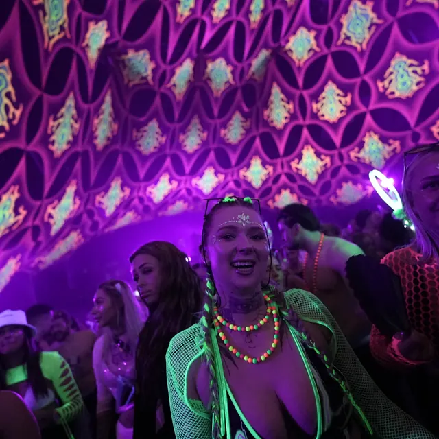 Festivalgæst med grønne fletninger og neonhalskæder under mønstrede loftsdekorationer.