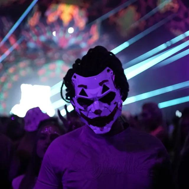 Festivalgæst med geometrisk maske foran blå laserstråler og farvede dekorationer.