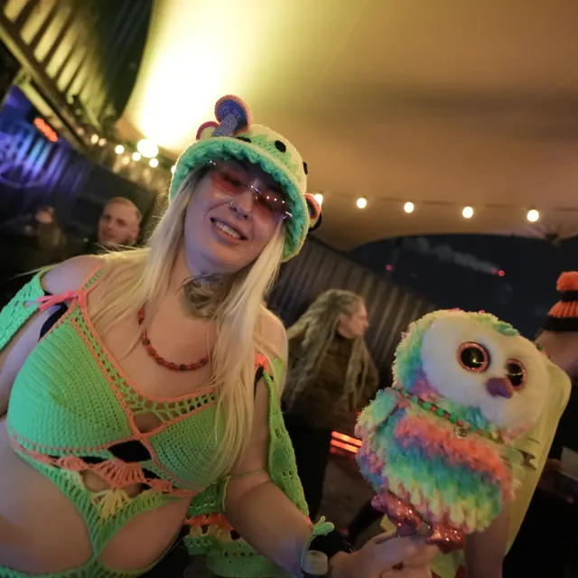 Festivalgæst med en farverig hat og en plysugle under en lyskæde.