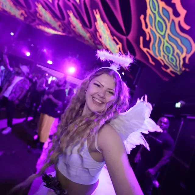 Smilende festivalgæst med hvide englevinger og glorie i lilla lys.