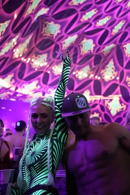 To festivalgæster under farverige dekorationer, den ene i grønne spiralmønstre med armen løftet.