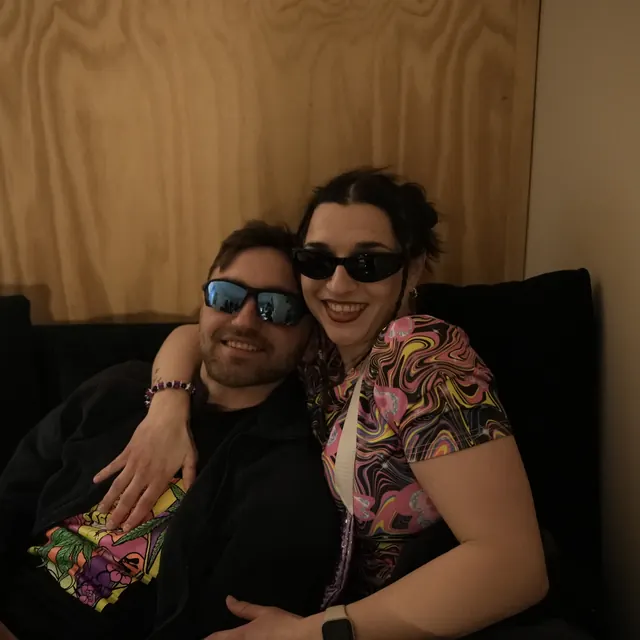 To smilende gæster med solbriller sidder sammen på en sofa.